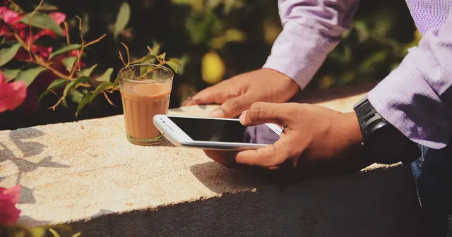 Mãos segurando um celular ao lado de um copo de café numa mesa de pedra, capa do guia de pix no delivery